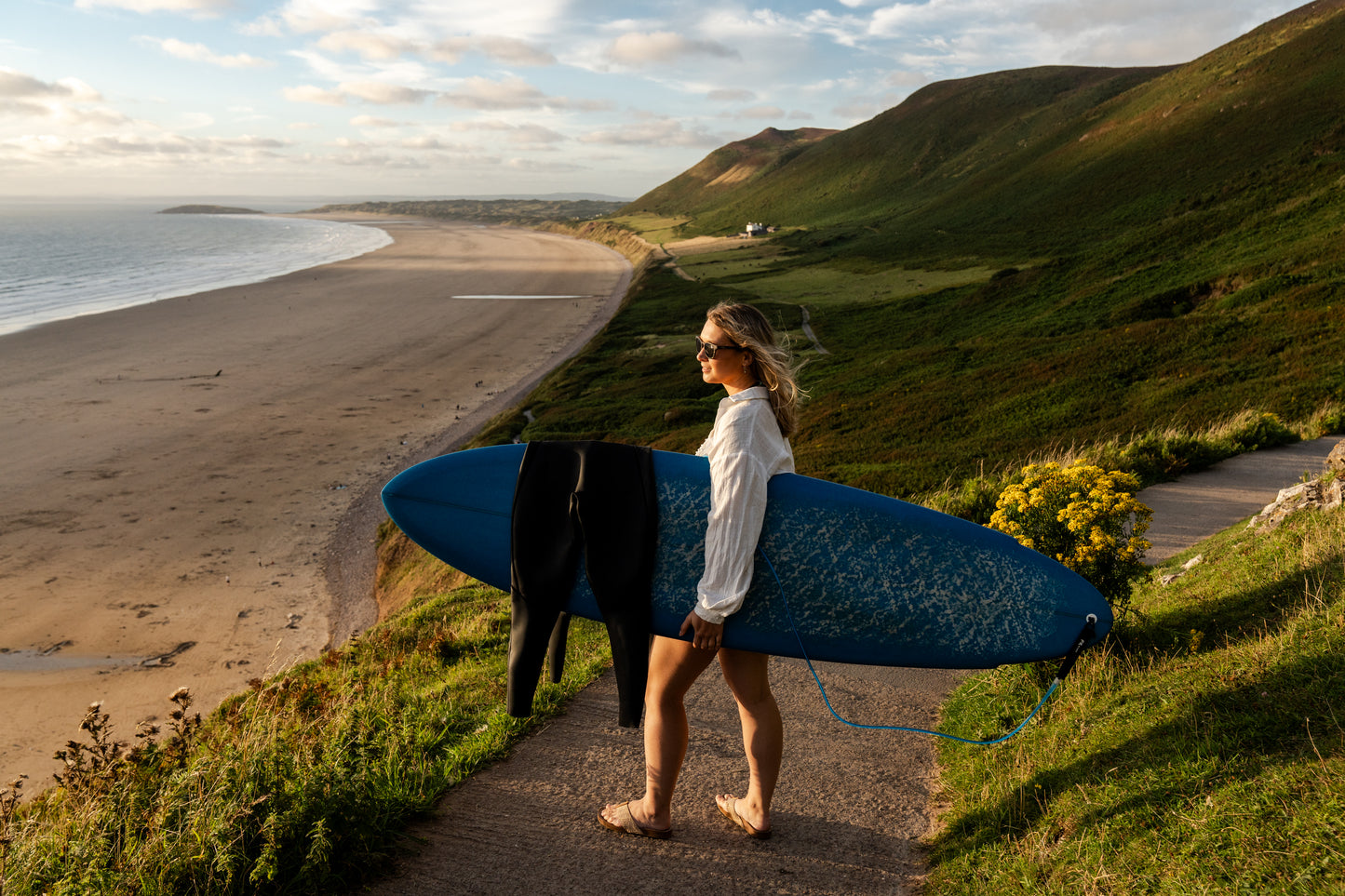 Surfer hold surfboard on hills over Welsh coastline.