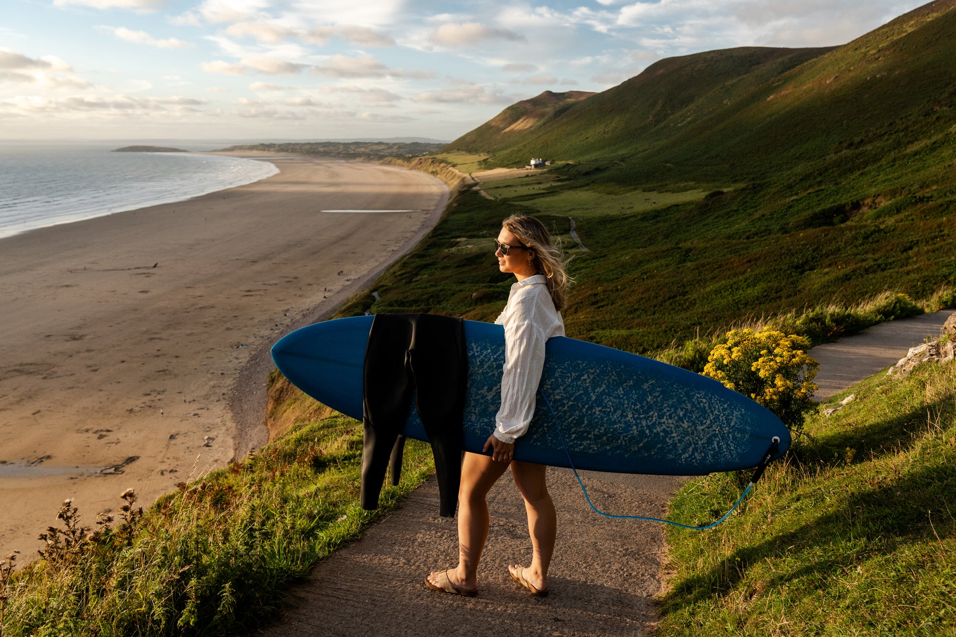 Surfer hold surfboard on hills over Welsh coastline.