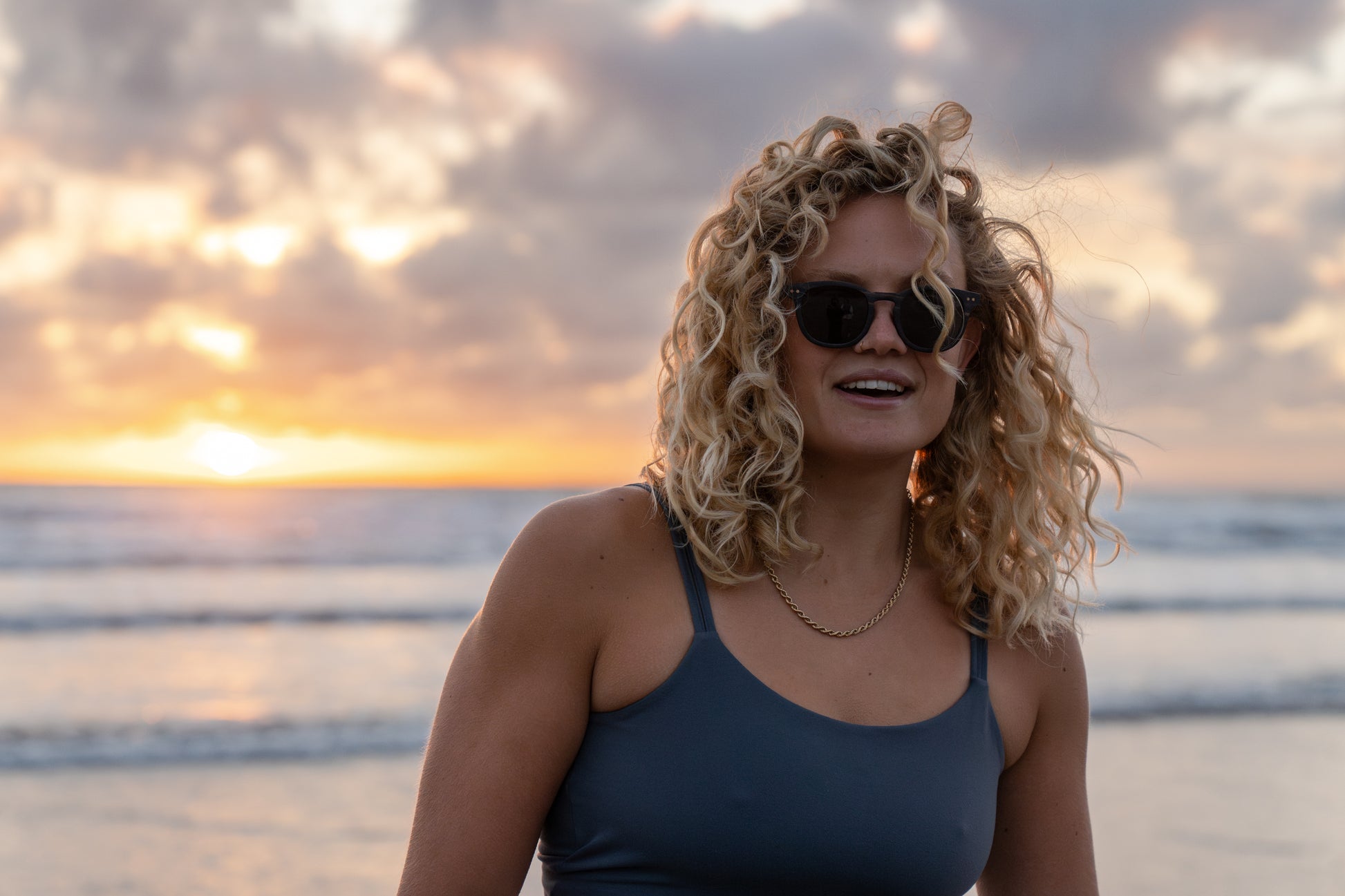 A person wearing the Shaka Black-Daughters of the Sea Limited Edition sunglasses, smiling at the beach during sunset.