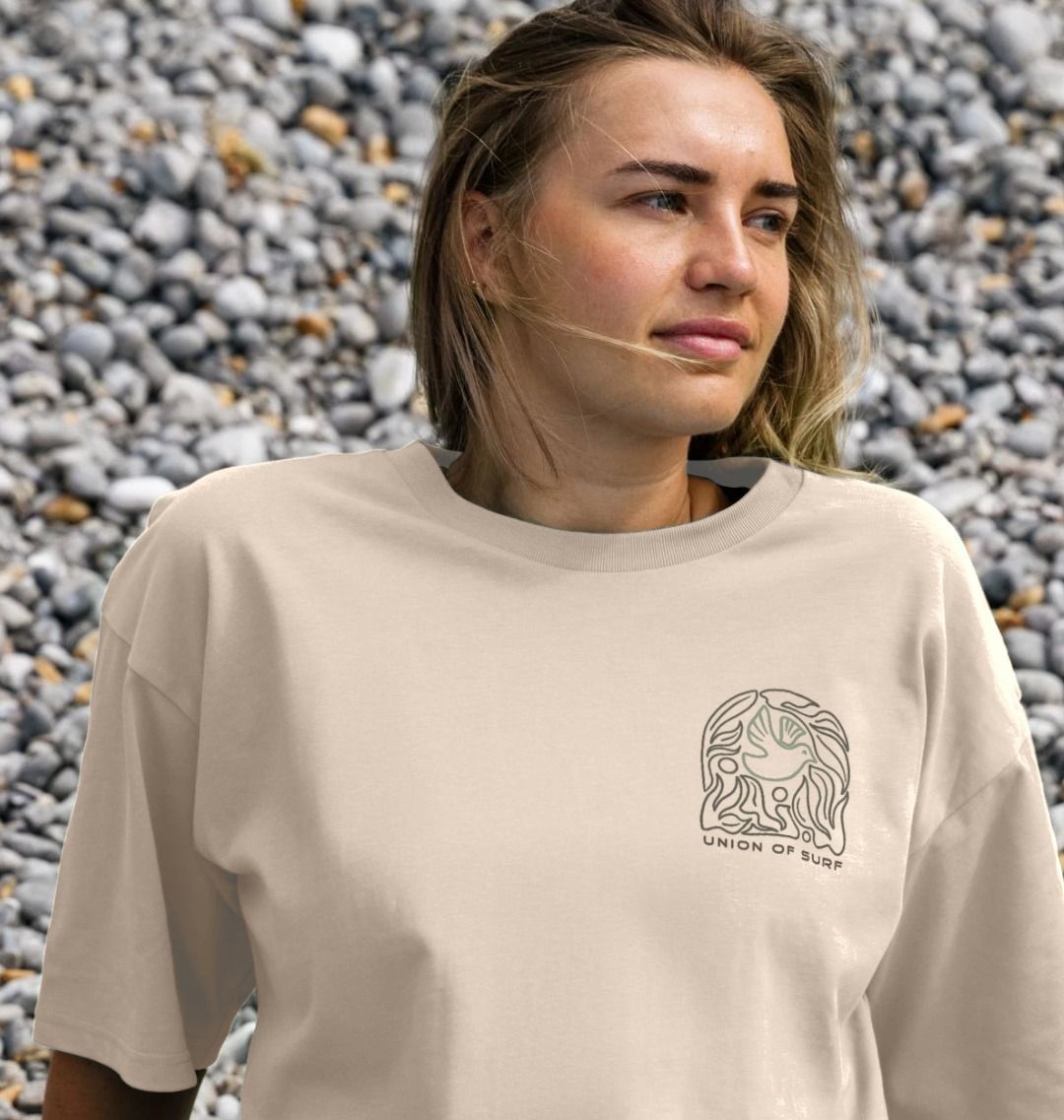 Woman wearing a beige t-shirt with a logo and text, standing against a stone wall.