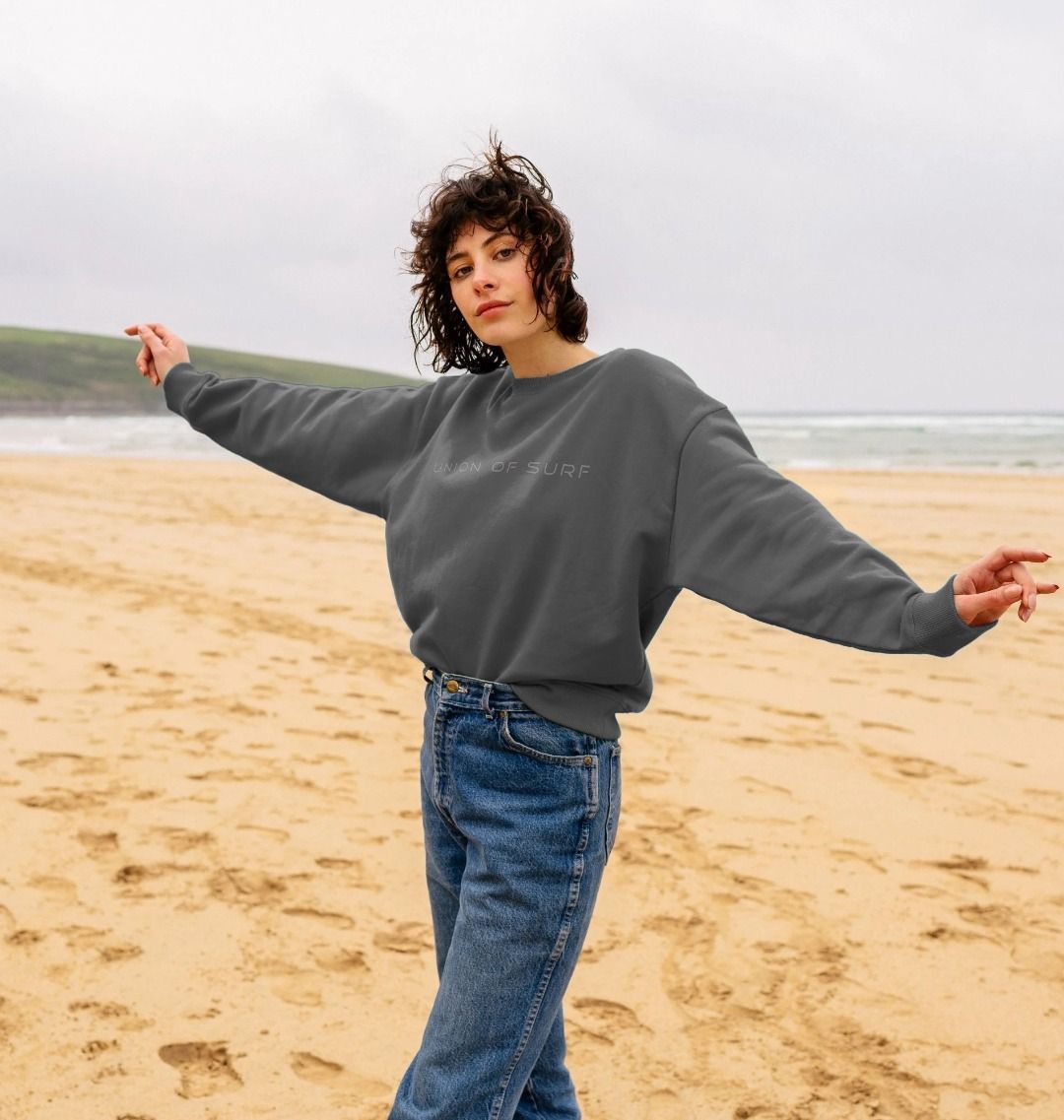 A woman standing on a beach wearing a gray Union Soft Touch Sweater and blue jeans, with the ocean in the background.
