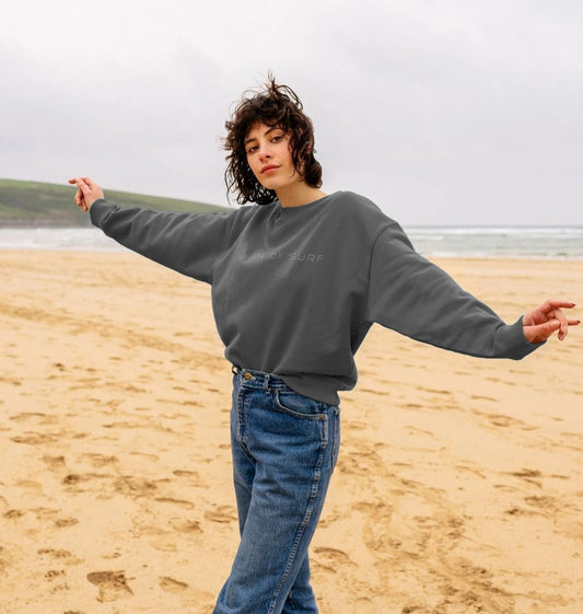 A woman standing on a beach wearing a gray Union Soft Touch Sweater and blue jeans, with the ocean in the background.