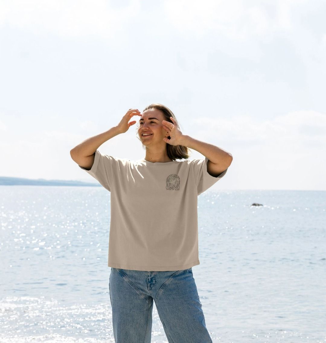 Person wearing a beige t-shirt and blue jeans standing on a beach with ocean in the background