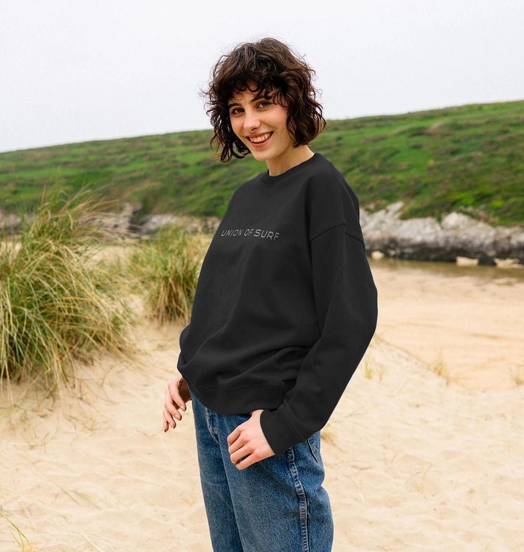 women stands on the beach wearing a relaxed fit sweater with union of surf on the front