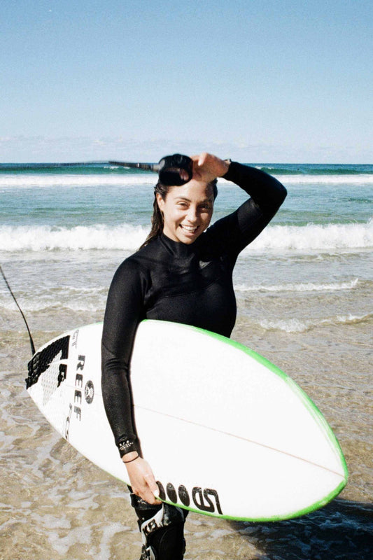 Woman in black full wetsuit holding white surfboard smiling at the beach UK Wetsuit Thickness & Temperature Guide: Find the Right Wetsuit for Any Water Condition