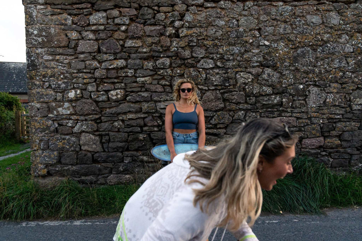 Woman wearing Shaka Black sunglasses and blue top standing by a stone wall with a surfboard.