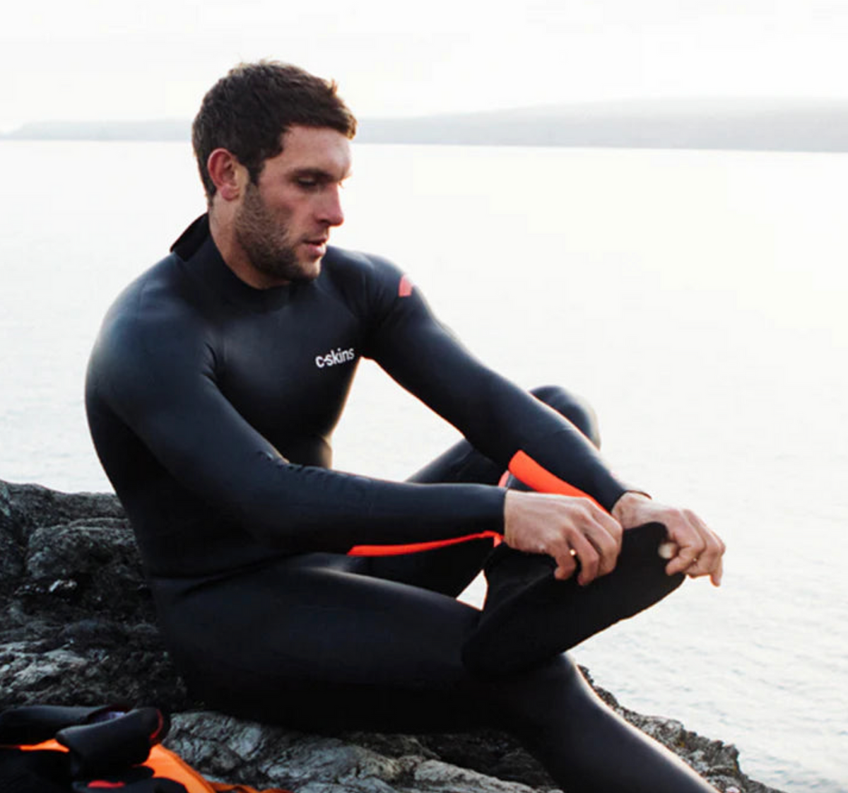 Black neoprene swim socks with orange accents worn by a man adjusting them, part of C Skins Swim Research gear.