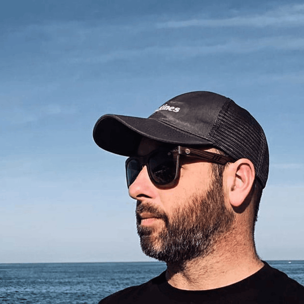 Man wearing Pit Dog sunglasses and a black cap against a blue ocean background.